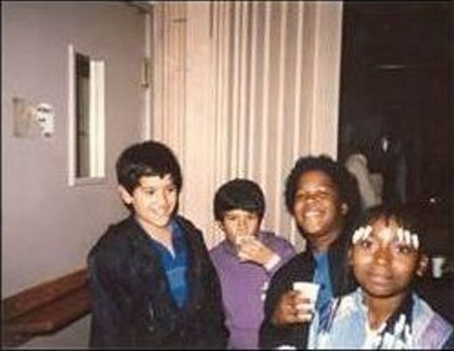 Four children indoors in the 1980s, one drinking from a cup