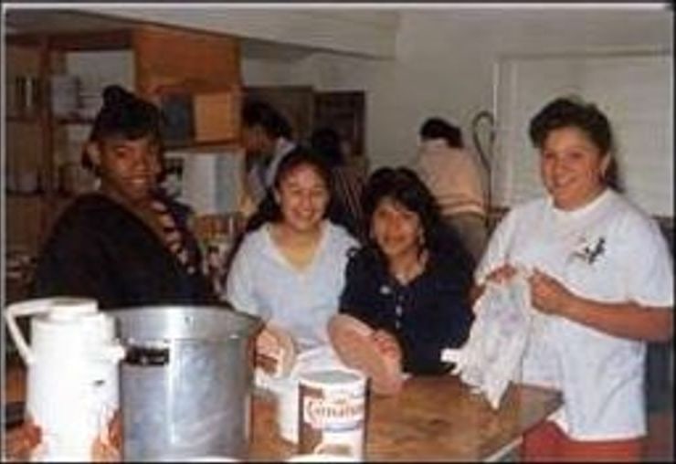 Four young women smiling behind a kitchen counter in the 1990s
