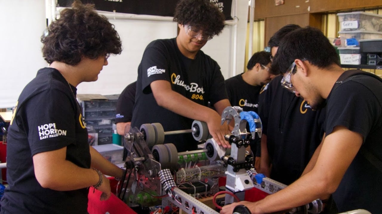 Churrobots students in team and Hope Horizon shirts assemble a robot chassis together in the workshop