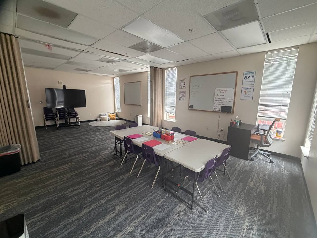 The classroom today: folding tables, stacked chairs against the wall, a rolled rug and a television on a stand