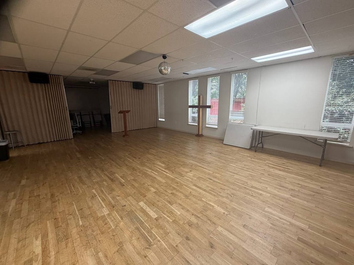 The gathering room today: an empty wood floor, a wooden cross, a lectern and a folding table against the wall