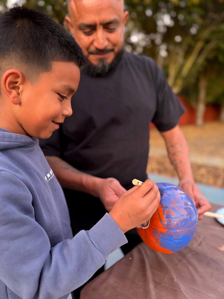 A boy paints a pumpkin blue while an adult holds it steady