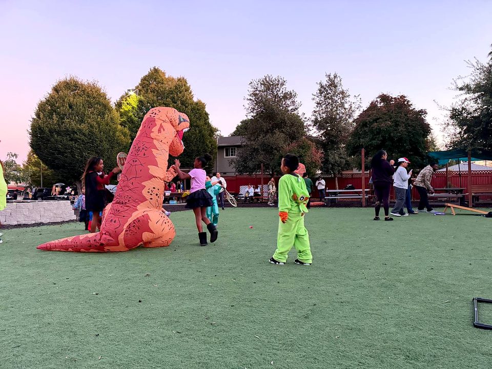 Families on the lawn at dusk, with two people in inflatable dinosaur costumes and a child in a bright green costume