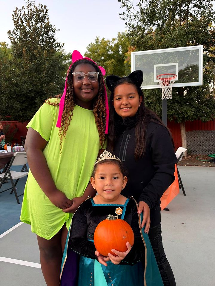 Three friends in costumes on the basketball court, the youngest in a princess dress holding a small pumpkin