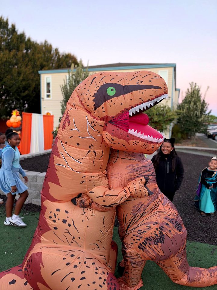 Two people in inflatable dinosaur costumes hug on the lawn while children laugh nearby