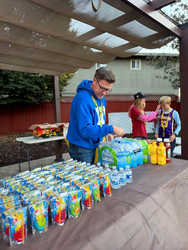 A volunteer in a blue hoodie sets out rows of juice pouches and water bottles under the pergola