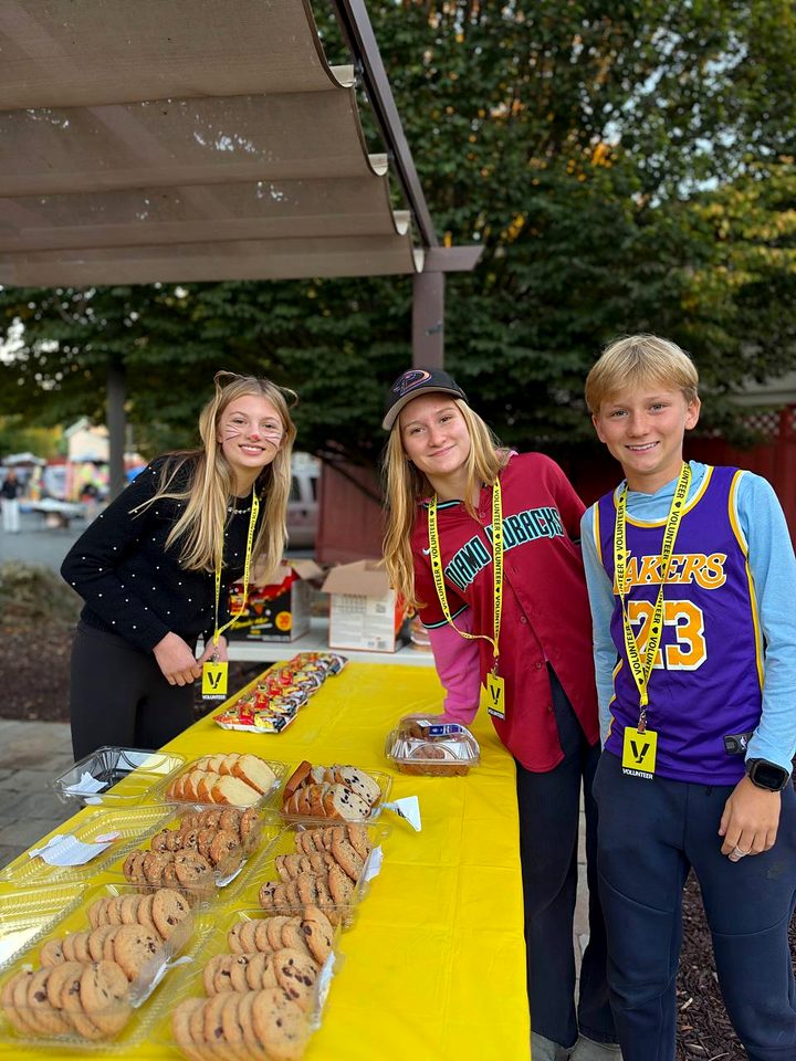 Three young volunteers in yellow lanyards stand behind a table of cookies