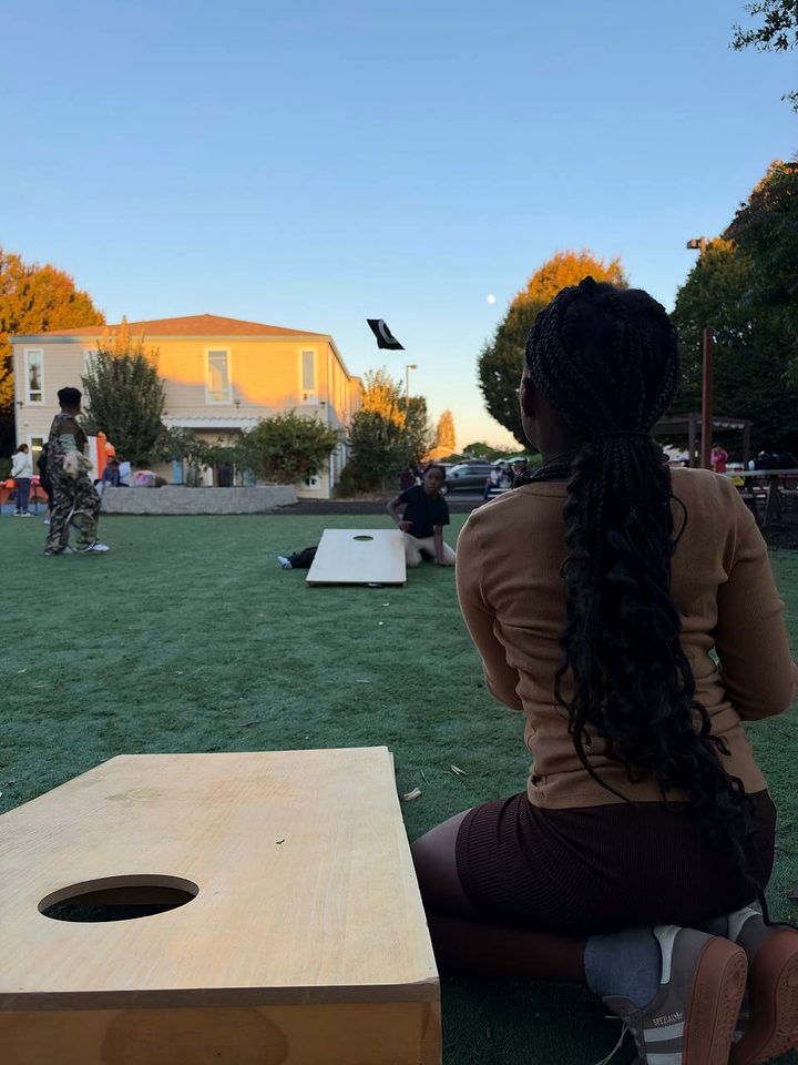 A person kneels on the lawn with a beanbag behind a cornhole board as the sun sets