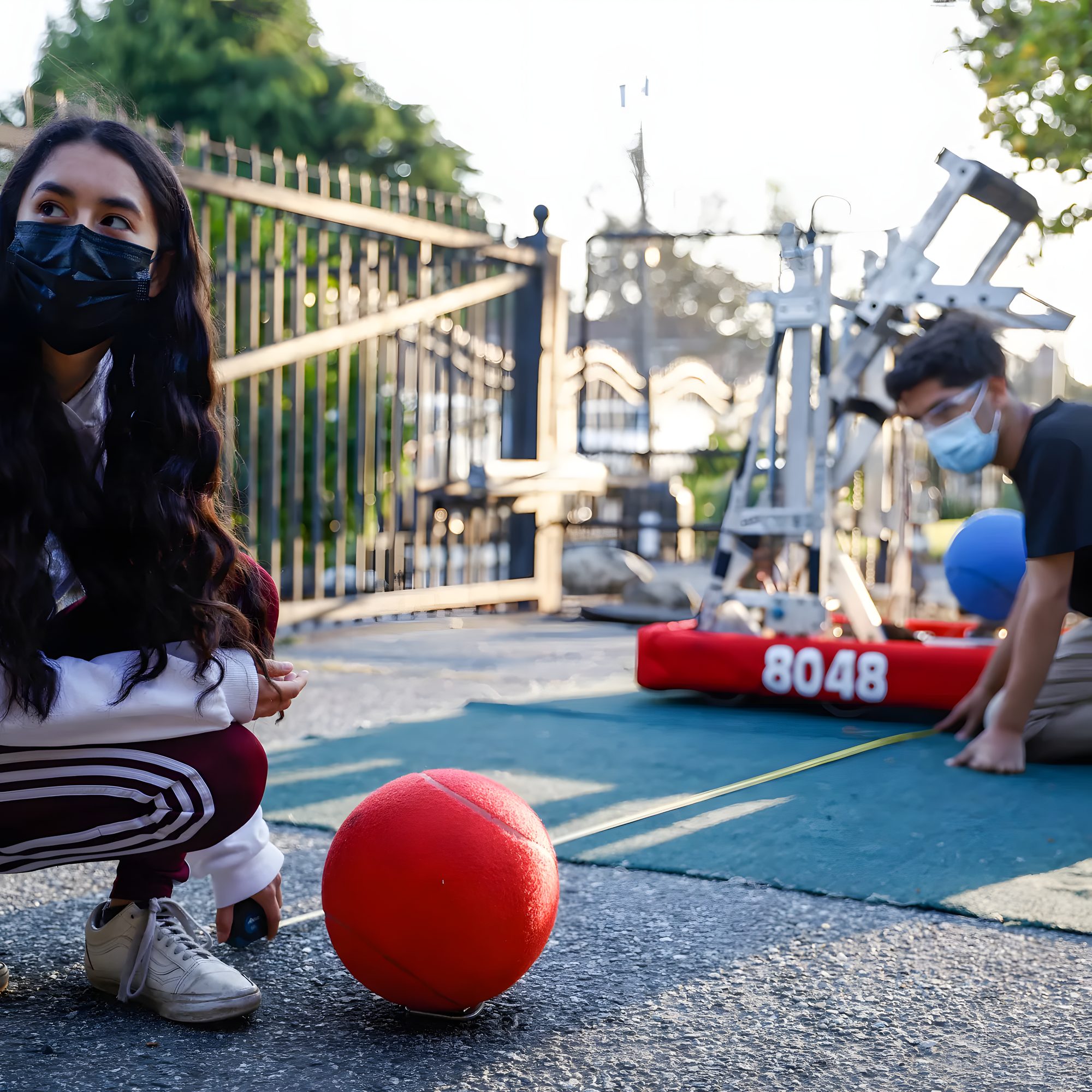 A student kneels beside the robot on an outdoor practice field, a red ball in the foreground